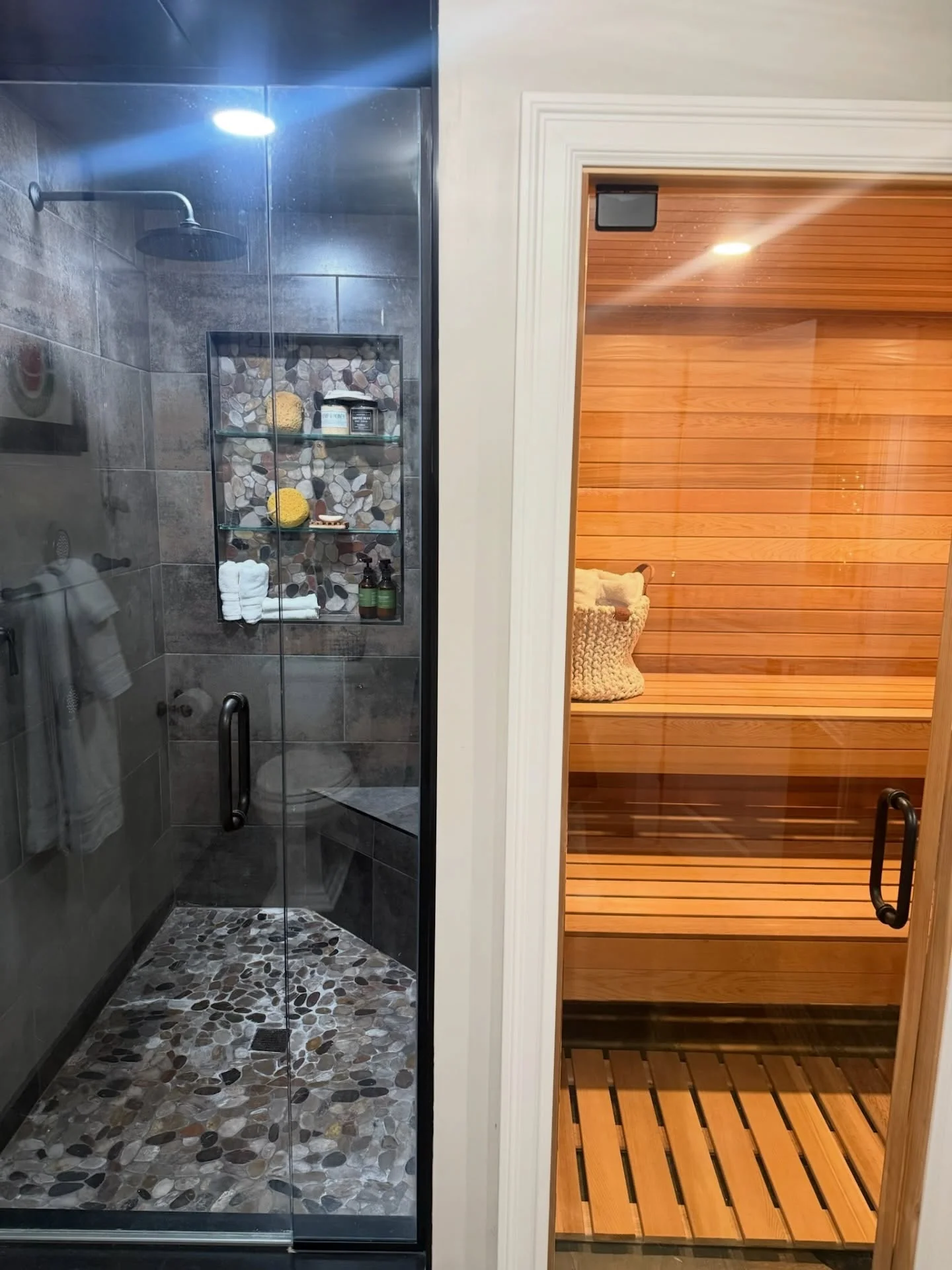Dark tile shower with river rock floor and glass niche shelving next to cedar sauna with glass door