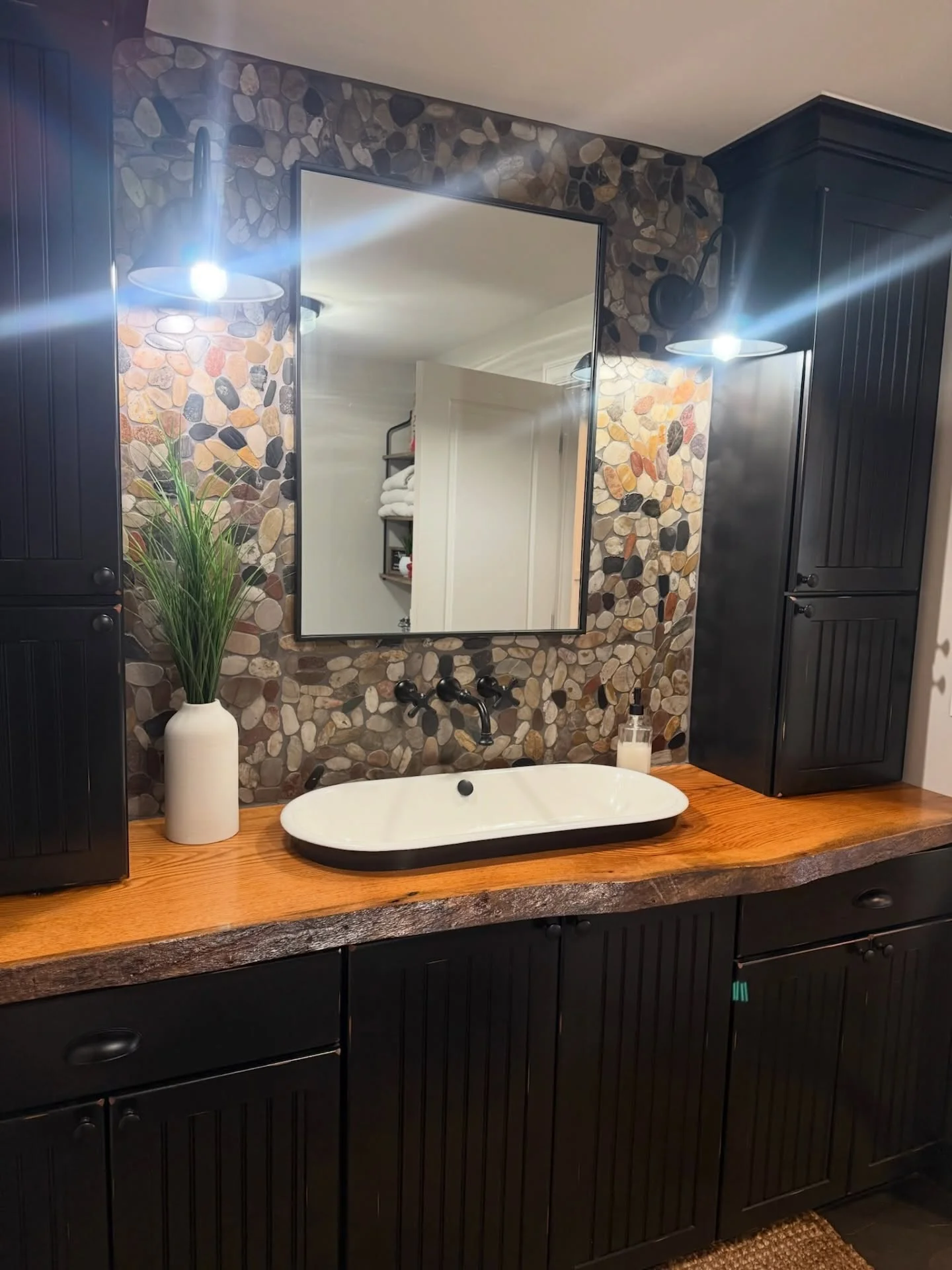 Black vanity with wood countertop, pebble stone accent wall, dual mirrors with LED lighting