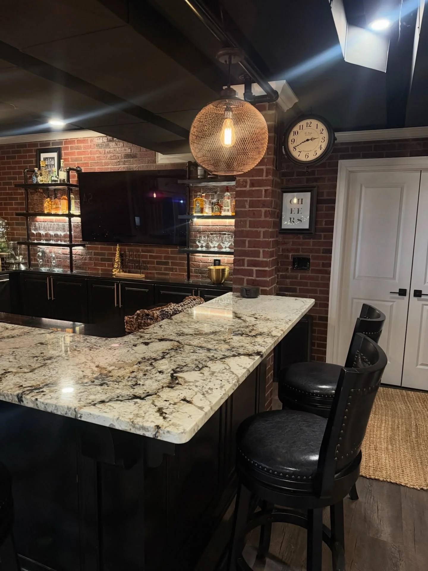Basement bar with granite countertop, black cabinetry, exposed brick wall, and custom shelving